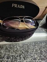 Photo of PRADA aviator sunglasses resting in the black Prada branded hard case, showing gradient lenses from purple to yellow, gold-tone metal frame, and Prada logo on lens.