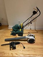 Green electric lawn mower with four wheels and handle, Ryobi electric blower with tube, and black McCulloch chainsaw attachment lying on wooden floor.