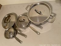 Four stainless steel pots and pans with three lids arranged on a flat surface.