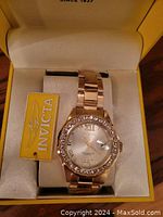 Front angle of Invicta watch showing gold tone bracelet, crystal bezel, dial with Roman numerals and date window, inside yellow Invicta box