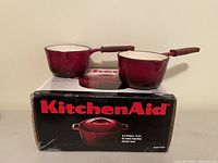 Two KitchenAid enamel cast iron sauce pots in deep red color with cream interior, placed on top of a boxed KitchenAid Dutch oven.