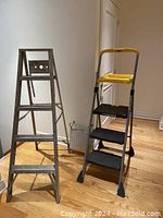 Two ladders standing upright on a wood floor including an aluminum step ladder and a gray and yellow plastic Cosco step ladder.