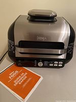 Front and side view of Ninja Foodi air fryer showing stainless steel and black plastic body with control panel and recipe guide booklet beside it.