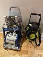 Two machines side by side on hardwood floor: blue/white AquaVac on dolly (left) and green/black Power It! unit (right)