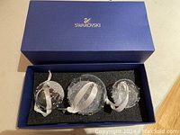 Three Swarovski spherical crystal ornaments in blue box with foam insert, showing size variation and hanging ribbons.