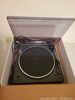 Black Audio-Technica USB turntable with open clear dust cover inside shipping box, power and audio cables visible.