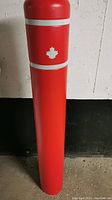 Full front view of red bollard cover with two white reflective stripes and maple leaf decoration near top.