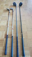 Four golf clubs including two irons and two woods, varying sizes and grips, laid out on parquet floor.