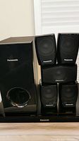Front view of five black Panasonic wall speakers and one black glossy subwoofer placed vertically on a wooden surface.