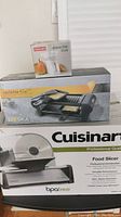 Boxed Cuisinart Food Slicer, Roscan Deluxe Diet Scale, and Boska Raclette Mini grill stacked for sale.