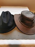 Two hats side by side on a light colored surface: a black wool hat and a brown leather hat with braided band.