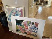 Three art pieces leaning against a wall near kitchen area. One art print shows outdoor scene with pink chairs, another partially visible framed abstract Kandinsky print with pastel tones, and a third not visible here.