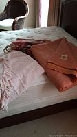 Three folded bedding items on a double mattress: a terracotta wool blanket with label, a pink and cream wool throw with fringes, and a pale pink cotton spread with fringes.