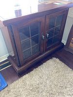 Front angled view of a dark wooden cabinet with two glass doors and visible shelving inside, placed on a wooden floor next to a carpet.