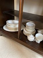 Photo of stacked dinnerware set showing plates, saucers, tea cups, sugar bowl, creamer jug, and serving platter arranged on a wooden shelf.