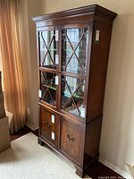 Full view of the wooden china cabinet showing upper glass doors with cross muntin design and lower wooden doors, standing on a carpeted floor near a curtain.