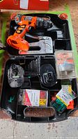 Complete set inside black plastic carry case on wooden surface. Visible cordless drill, batteries, Bostitcher, hole saws, drill bits, and plastic plugs.