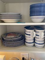 Shelved view of multiple dinner plates, salad plates with fruit motifs, bowls, and mugs with blue stripes.