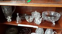 Photo of a wooden shelf with all the glassware items including a large cut glass bowl on the right, pitcher, bell, salt cellar, milk and sugar containers spread across the shelves.