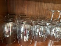 Photo of multiple clear stemless round liquor glasses stacked inside each other with some stemmed wine glasses visible at the back on a wood shelf.