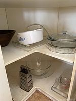 Photo of a corner shelf holding various kitchen items including a white CorningWare casserole dish with blue floral pattern, two clear glass casserole dishes with lids, a square glass baking dish, a metal box grater, and three clear glass bowls nesting inside each other.