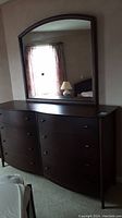 Front view of large dark brown wooden dresser with eight drawers and attached mirror reflecting room elements.