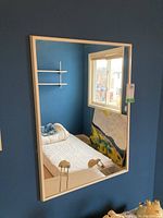 Mirror hanging on blue wall reflecting a window, bed, shelf, and painting, showing frame color and mirror condition.