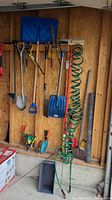 Photo showing hanging garden tools including shovels, rakes, broom, green coiled garden hose and metal rod on wooden wall
