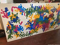 Front view of the large abstract painting on canvas resting against a wall, showing interconnected colorful shapes on white background.