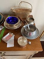 Group of serving items on wooden surface: wire basket, cobalt bowl, silver pedestal bowl, rectangular dish, silver mug, round container, sauce pan set.
