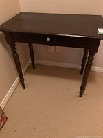 Front view of vintage wooden table showing single drawer and four turned legs against beige wall and carpeted floor.