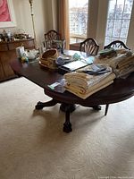 Full view of vintage wood dining table with leaves extended, dark finish, set in room with carpet and other furniture. Various items on top of table.