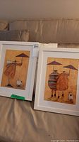 Two framed wall art prints shown resting on a beige couch, displayed side-by-side, each with white frames and featuring orange-brown stylized artwork with red umbrellas and small animals.