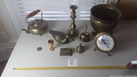 Full view of all brass items arranged on white surface next to ruler for scale, showing kettle, duck, candlestick, clock, flower pot, bell, small cup, and box.