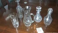 Wide view showing four crystal decanters and one glass pitcher on wooden table.