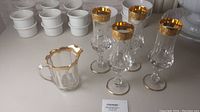 Set of four clear Murano Medici style wine glasses with ornate gold rims, alongside a small glass pitcher with a scalloped gold rim and handle.