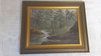 Framed oil painting of wooded stream landscape shown fully in its frame with fabric backdrop.