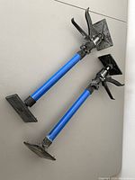 Two blue and black adjustable cabinet helping hand jacks lying horizontally, showing square gripping plates and trigger mechanisms on top ends.