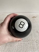 Hand holding classic black Magic 8 Ball toy showing the white circle with the number 8 on the surface, minor surface imperfections visible.