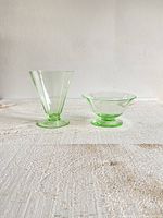 Two vintage green uranium glass dishes sitting on a textured surface against a plain white wall under natural light. Shows the taller conical dish and the shorter octagonal-rim dish.