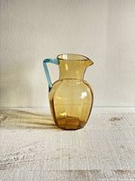 Smokey yellow brown glass pitcher with blue handle on a white textured surface against a white wall.