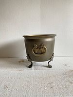 Full view of the round dark brass planter pot showing three footed legs and one side handle, sitting on a textured surface against neutral wall.