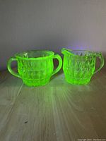 Photo of both vintage green Depression glass creamer and sugar bowl, glowing bright green under UV light, showing geometric diamond pattern and handles.