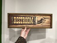 Vintage wooden restroom sign held up by a hand, showing full sign design and wood frame.