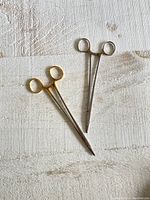 Two vintage metal medical suture clamps lying on a light textured surface, showing front view with finger ring handles and slim jaws.