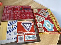 Photo showing the entire vintage Spirograph set including the red plastic tray with gears and frames, pens, instruction booklet, paper packs, and box.