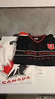Photo showing both black and white Team Canada hockey jerseys folded, with visible Team Canada logos and partial Bauer branding on white jersey.
