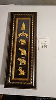 Front view of framed rectangular wall art with six gold-tone animal figurines including elephants and a camel on a black background, ornate dark wood frame with gold detailing