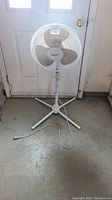 White Orbit floor fan standing on a concrete floor in front of a door. Shows fan in full, power cord extended on the floor.
