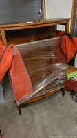 Wooden armoire wrapped in plastic with visible glass doors and wooden shelves inside.